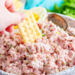 ham salad shown in a white speckled bowl with a hand dipping a club cracker into the ham salad