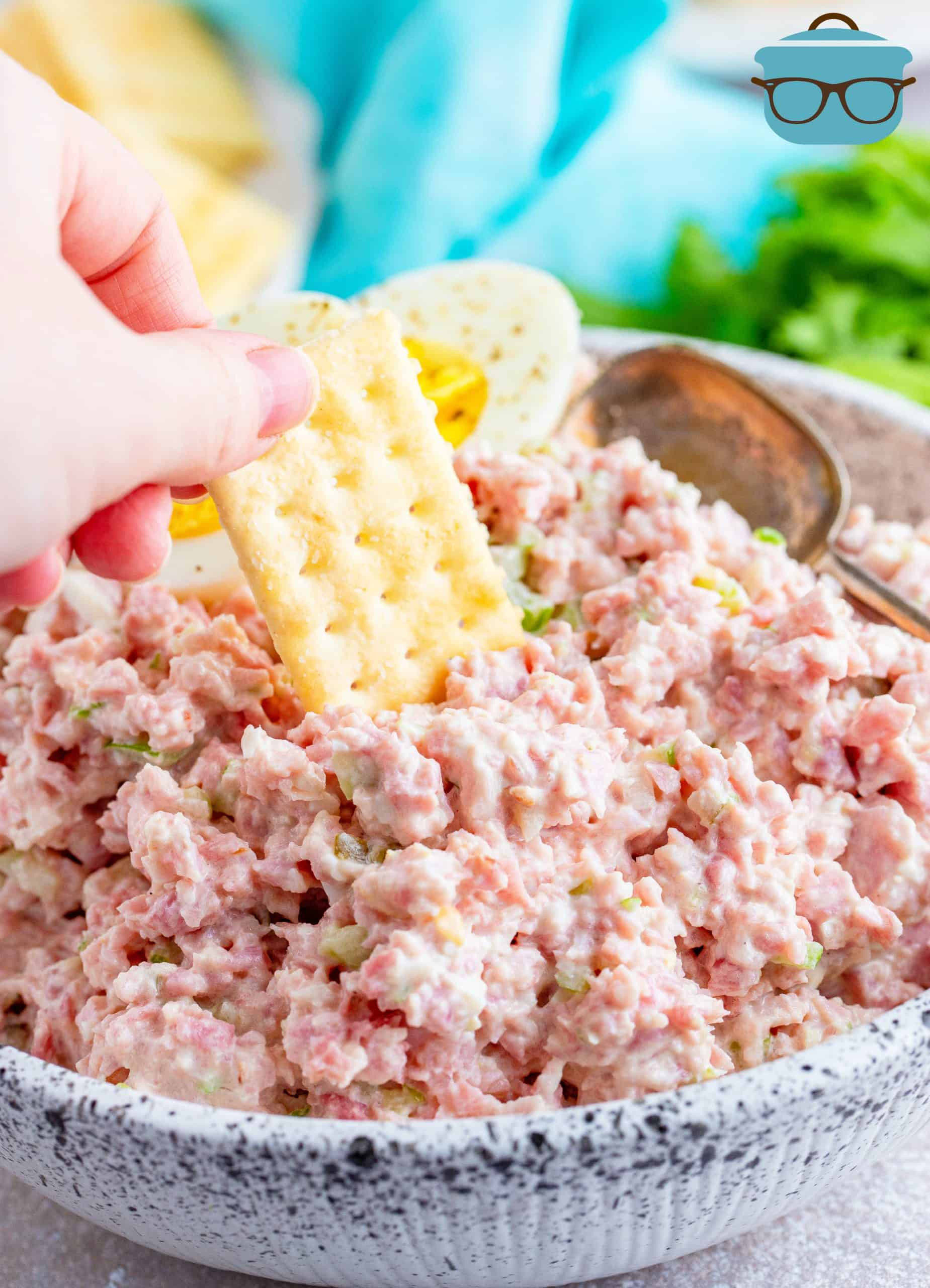 ham salad shown in a white speckled bowl with a hand dipping a club cracker into the ham salad