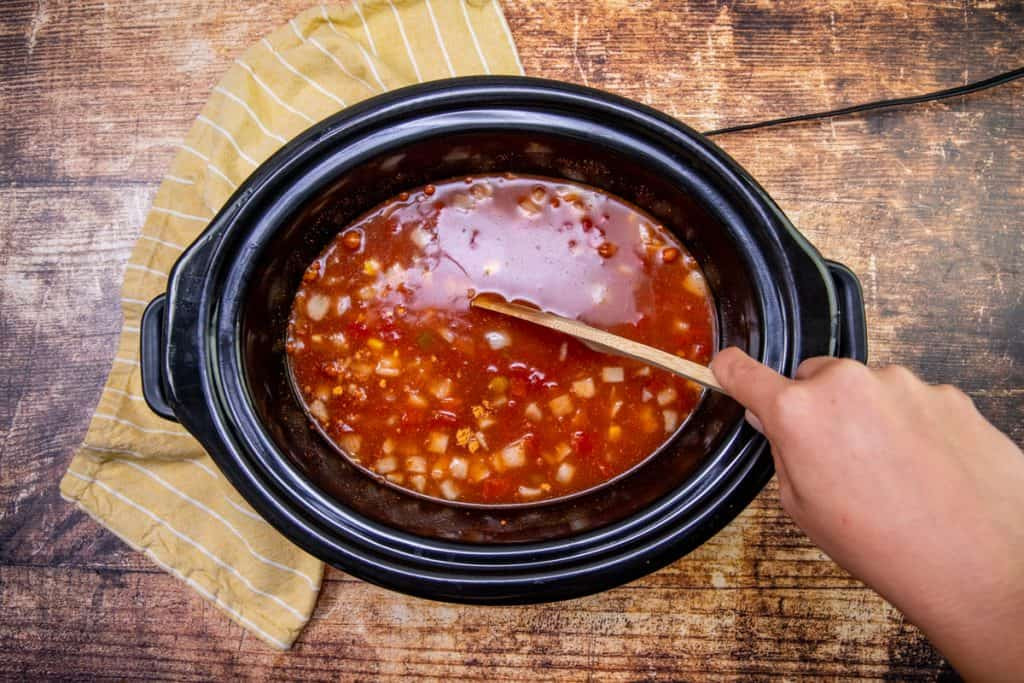 Raw chicken, enchilada sauce, seasoning, and diced onions being stirred in a crock pot, illustrating the initial steps of layering and combining ingredients