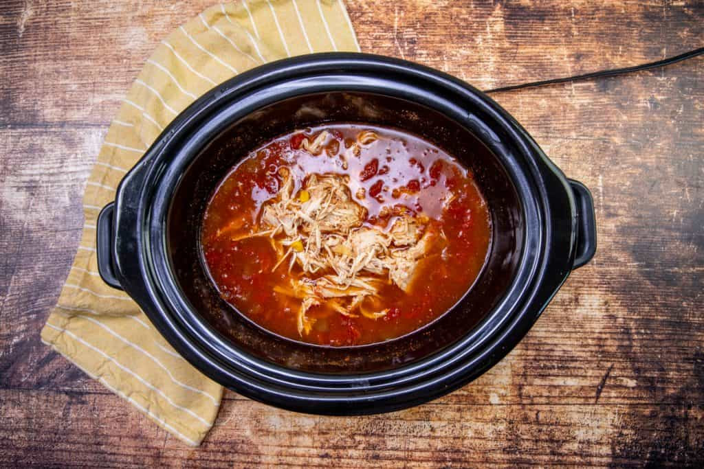 Shredded chicken being added back into the crock pot with the soup liquid, demonstrating the step of incorporating the shredded chicken before the final simmer