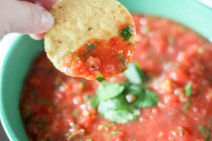 Overhead shot of a bowl of Pioneer Woman Restaurant Salsa with a spoon scooping some out, showcasing the chunky texture, alongside a basket of tortilla chips on a wooden table.