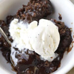 Close-up of chocolate cobbler in a white bowl, topped with a scoop of vanilla ice cream and a spoon resting in the bowl.