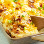 A close up shot of a wooden spoon serving twice baked potato casserole from a white baking dish.