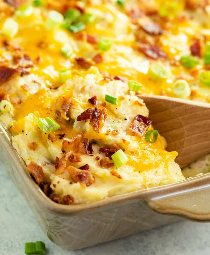 A close up shot of a wooden spoon serving twice baked potato casserole from a white baking dish.