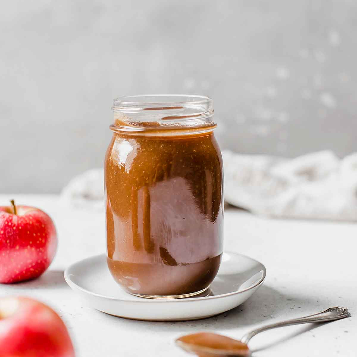 Image of a jar of apple butter on a white saucer, with a spoonful of apple butter in the foreground and two apples beside the jar.