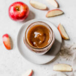 Overhead shot of a jar of apple butter on a white plate surrounded by sliced apples.