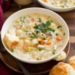 Bowls of creamy chicken pot pie soup garnished with fresh herbs and served with flaky biscuits on a rustic wooden table.