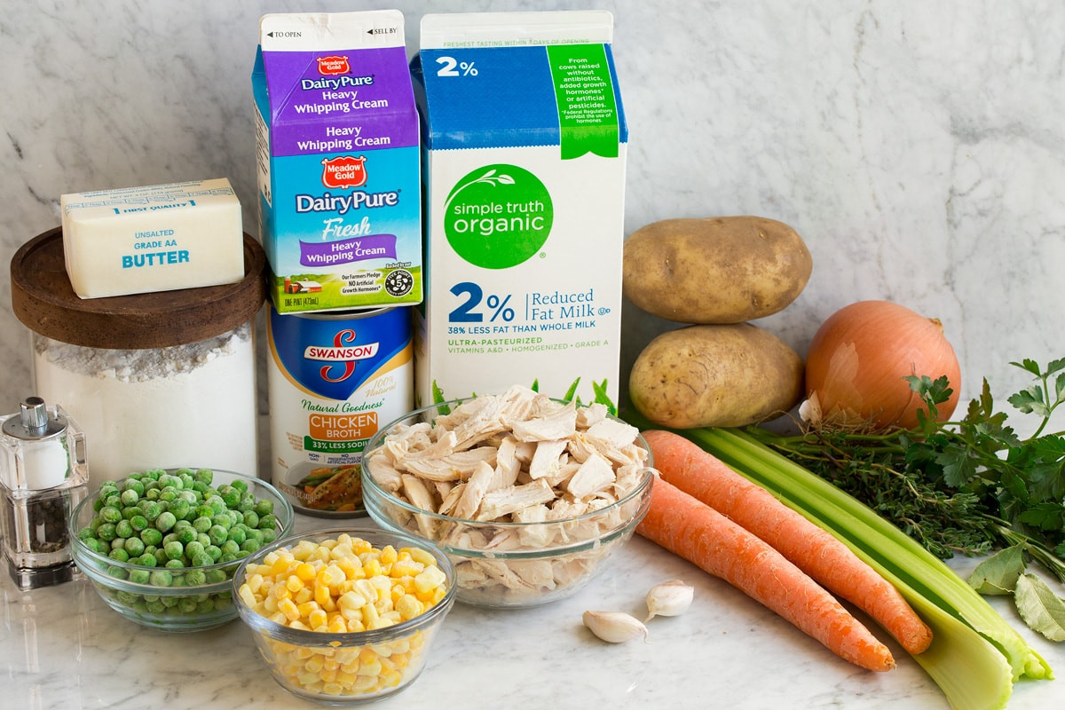 Ingredients for chicken pot pie soup laid out on a kitchen counter, including fresh vegetables, herbs, chicken broth, dairy, and cooked chicken.