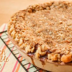 Close-up shot of a slice of peach crumble pie with a golden brown crust and crumble topping, served on a white plate.