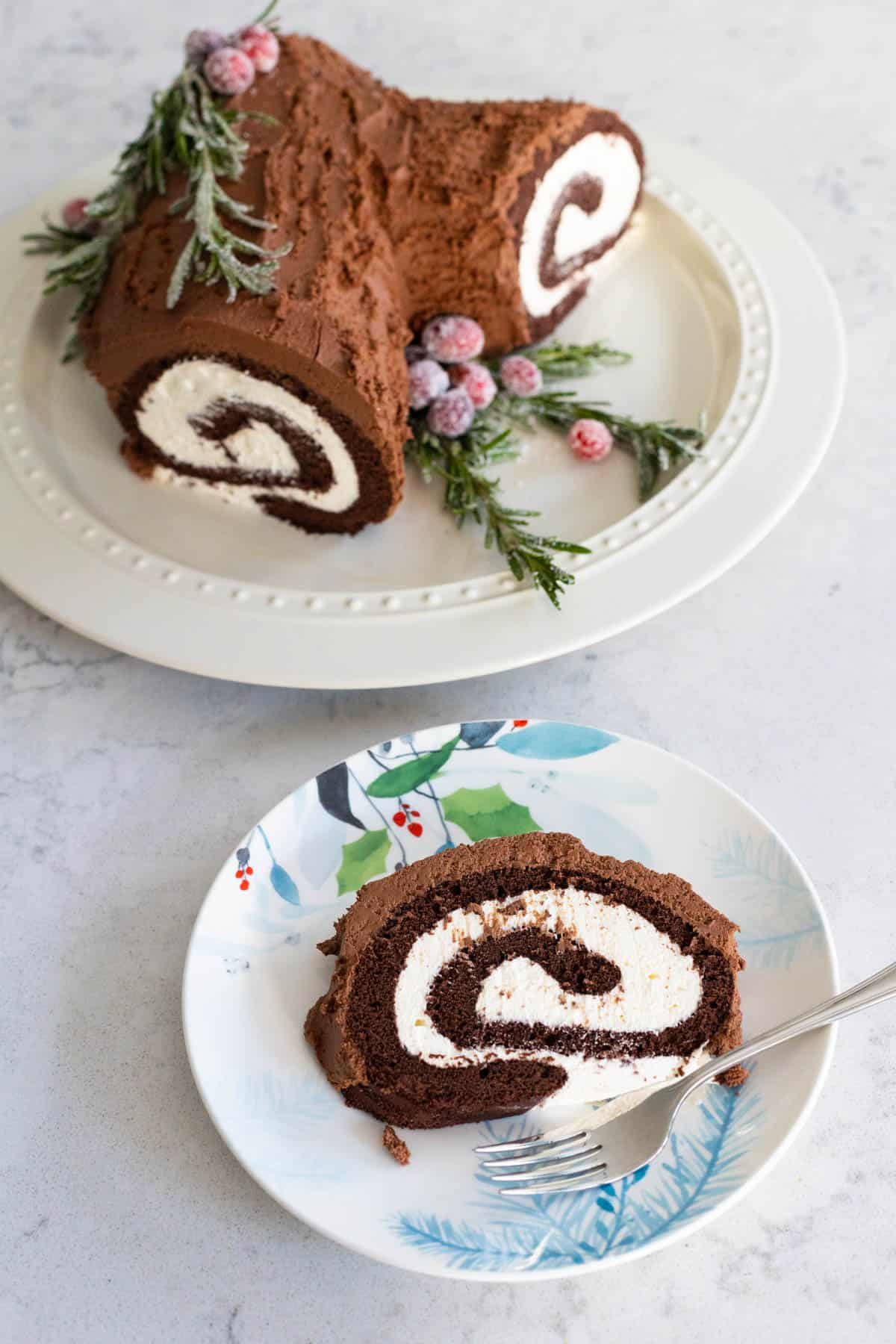 Sliced Pioneer Woman Yule Log Cake on a Christmas plate with a fork, with the whole cake on a platter in the background, showcasing the festive dessert.